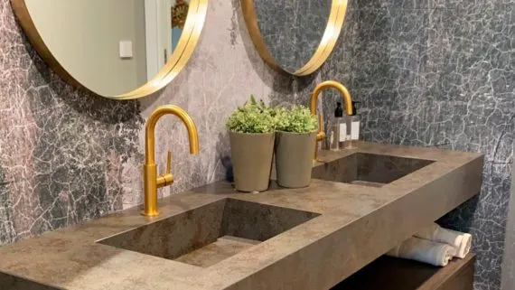 Lavabo de baño con encimera de piedra, dos grifos dorados y espejos circulares.