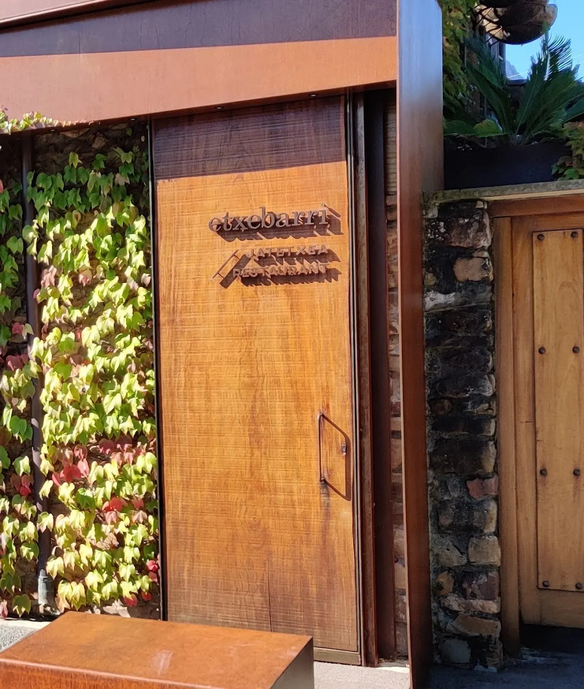 Entrada de Asador Etxebarri en Axpe, con puerta de madera y fachada cubierta de vegetación.