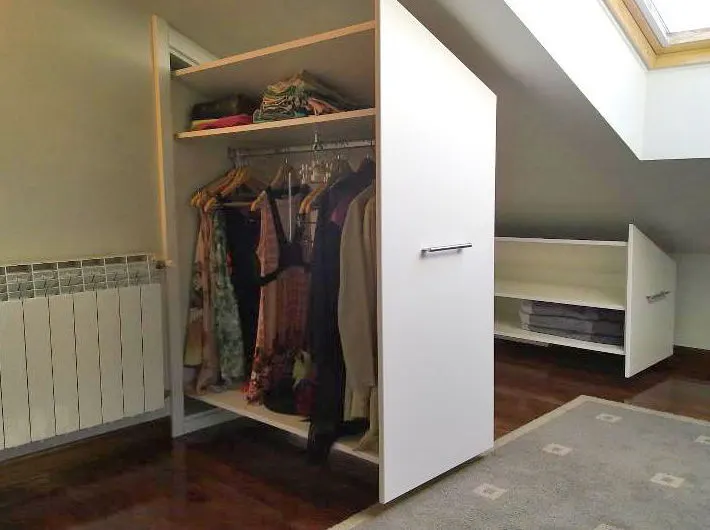 Armario empotrado bajo un techo inclinado, con ropa colgada y estantes