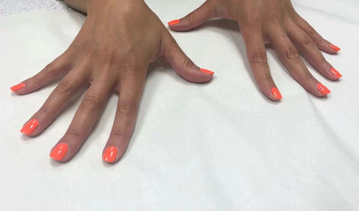 Manos con las uñas pintadas de coral sobre una superficie blanca.