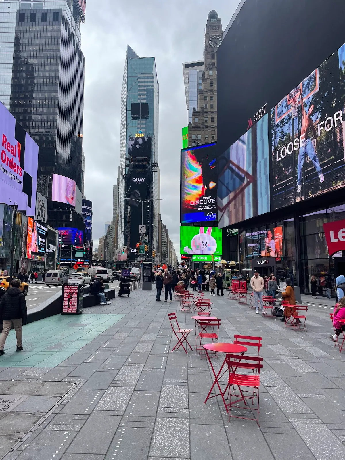 Vista de una plaza urbana con edificios altos, pantallas y mobiliario rojo.