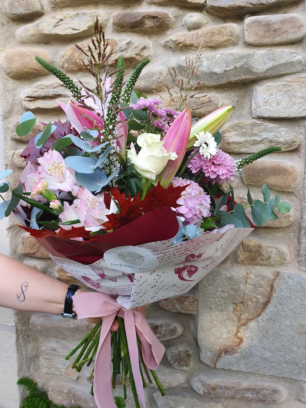 Ramo de flores variadas envuelto en papel junto a una pared de piedra
