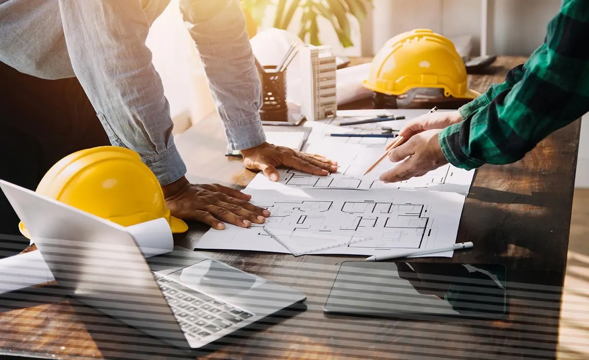 Personas revisan planos de una obra junto a cascos de seguridad y un ordenador.