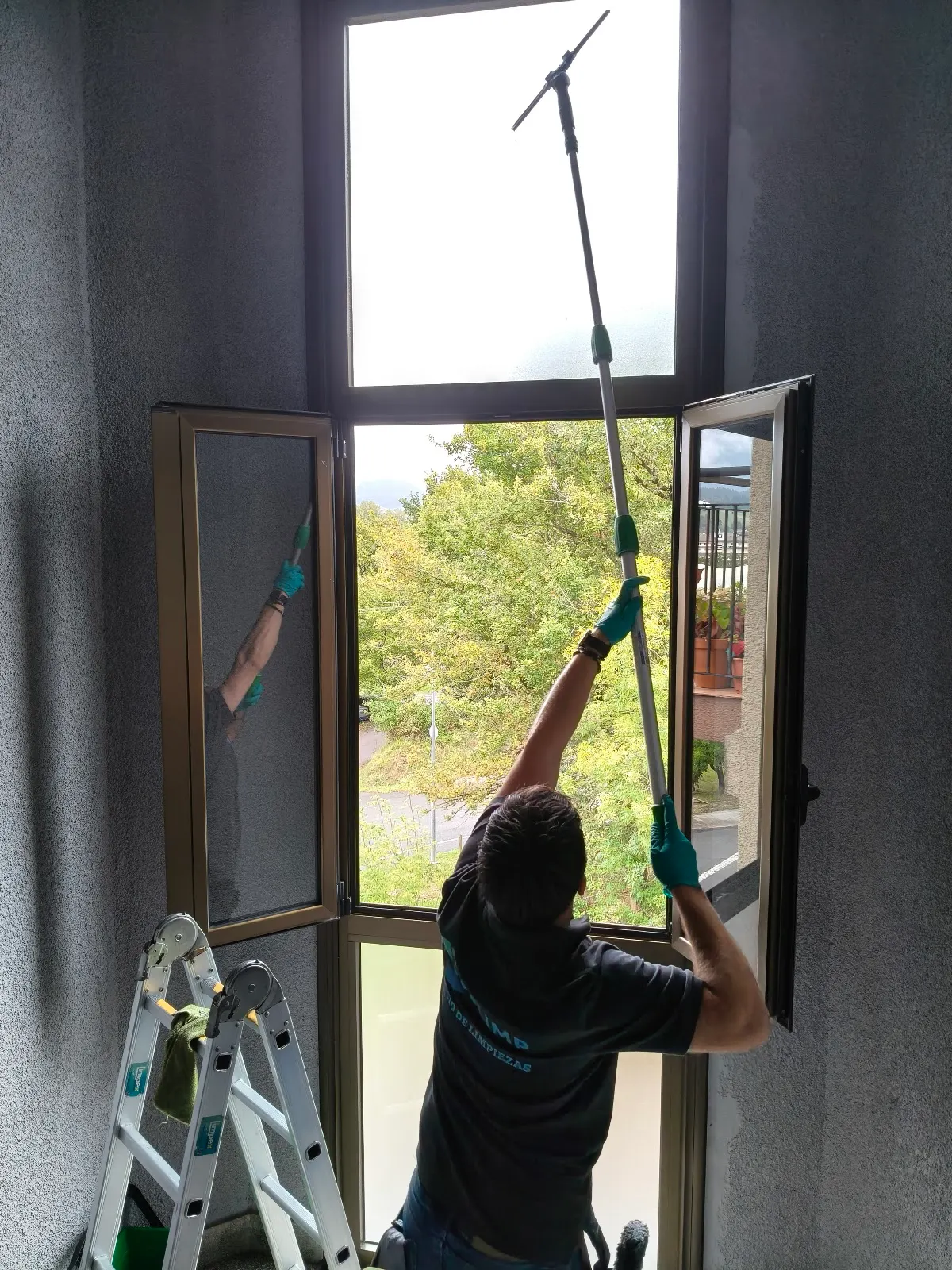 Una persona limpia una ventana con una herramienta extensible en un interior; Limpiezas COSLIMP GARBIKETAK.