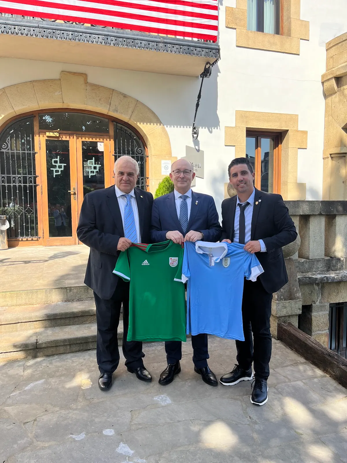 Tres hombres posan frente a un edificio con dos camisetas de fútbol; no se identifica el negocio.